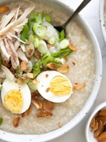 Bowl of chicken congee topped with shredded chicken, sliced boiled egg, lime wedges, green onions, and crispy shallots, with additional toppings in small bowls on the side.