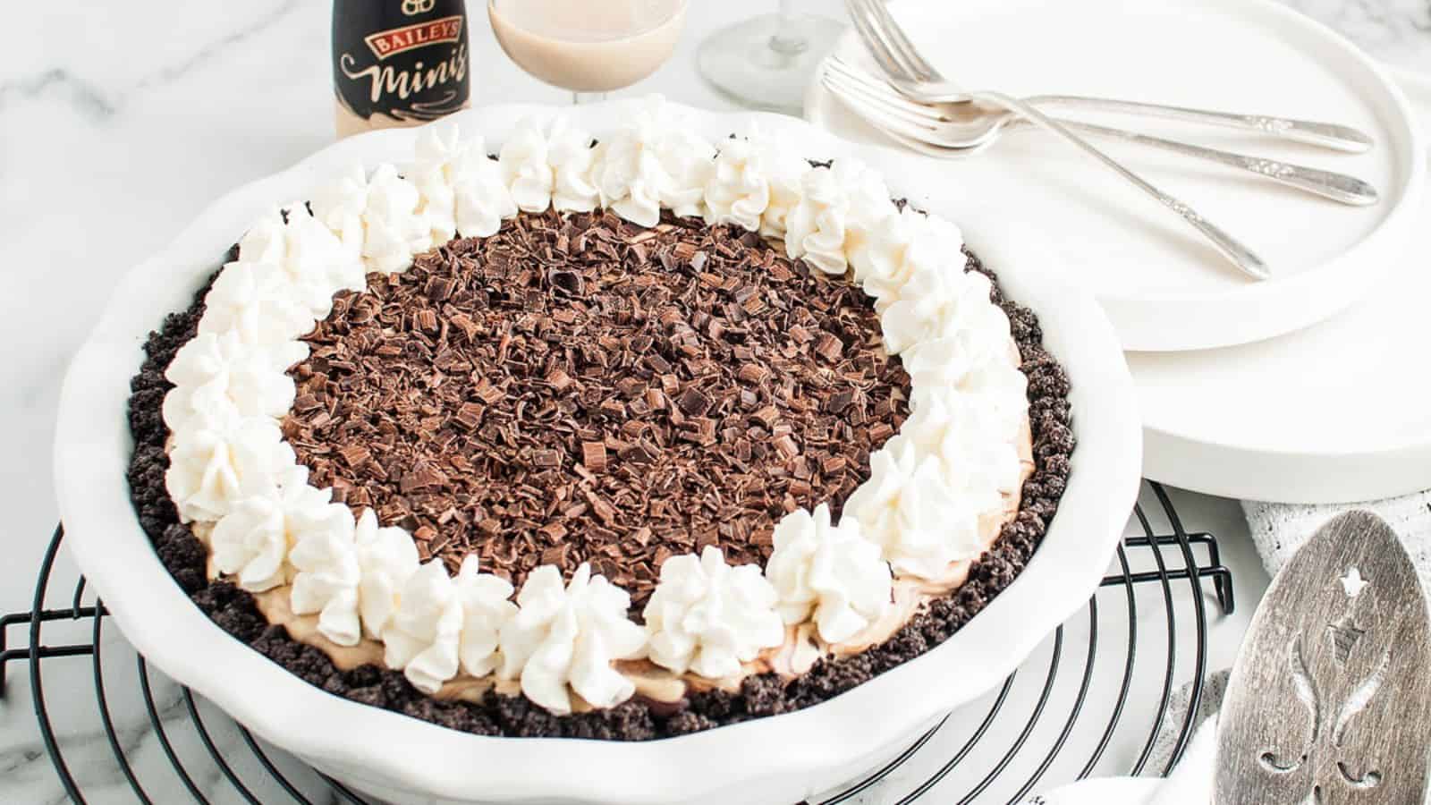 A chocolate cream pie topped with whipped cream and chocolate shavings in a white dish on a cooling rack, surrounded by dessert plates, forks, a serving spatula, and a Bailey's bottle in the background.