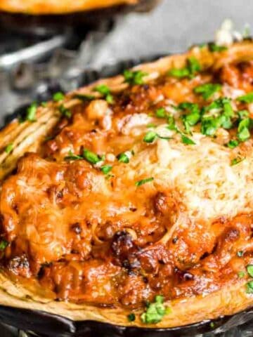 Baked eggplant filled with a savory mixture, topped with melted cheese and garnished with chopped parsley, on a textured surface.