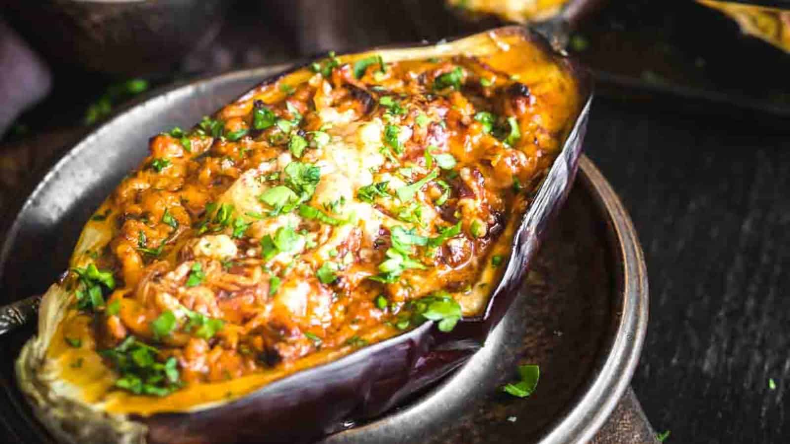 Stuffed eggplant with a baked, golden-brown filling, garnished with chopped herbs, on a dark plate.