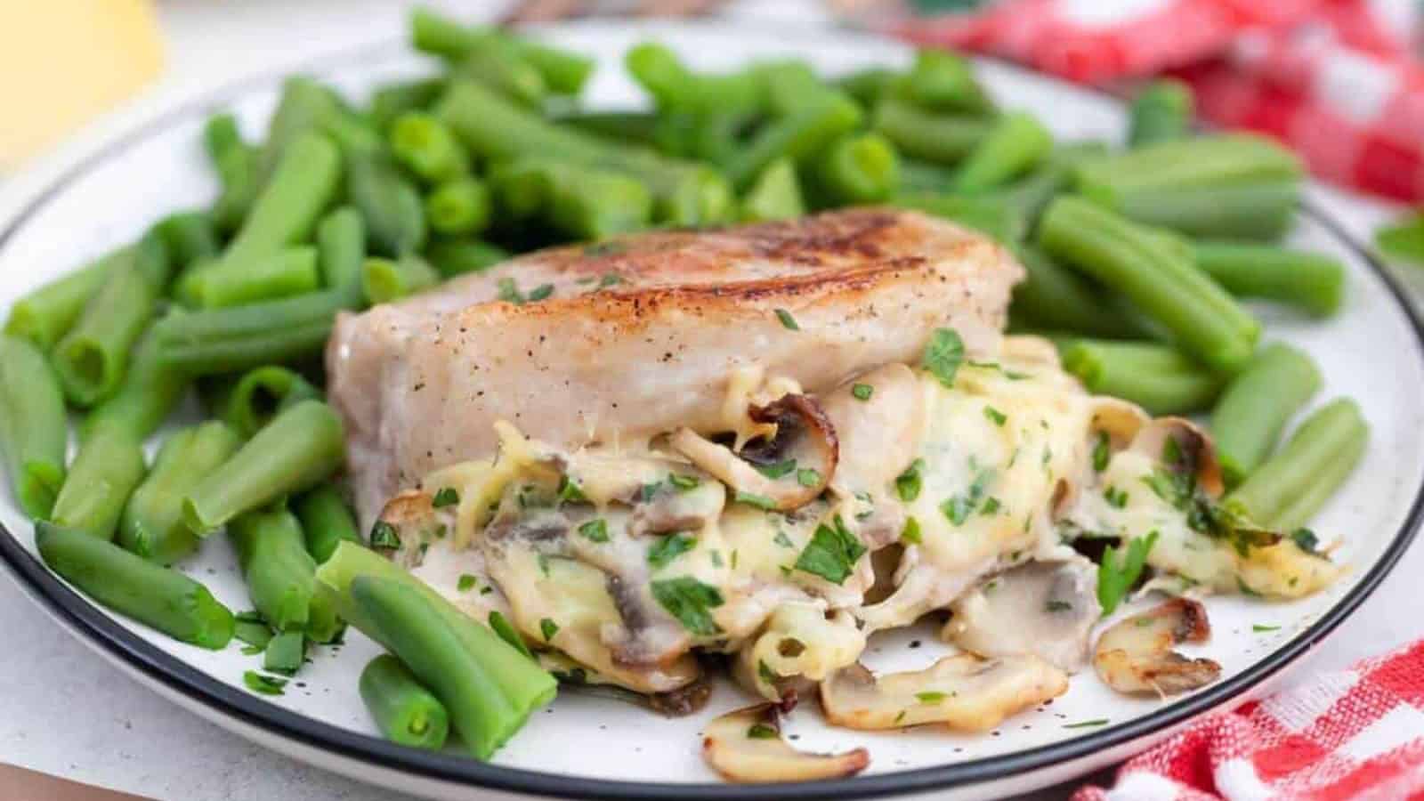 A plate with a stuffed pork chop topped with mushrooms and herbs, surrounded by steamed green beans.