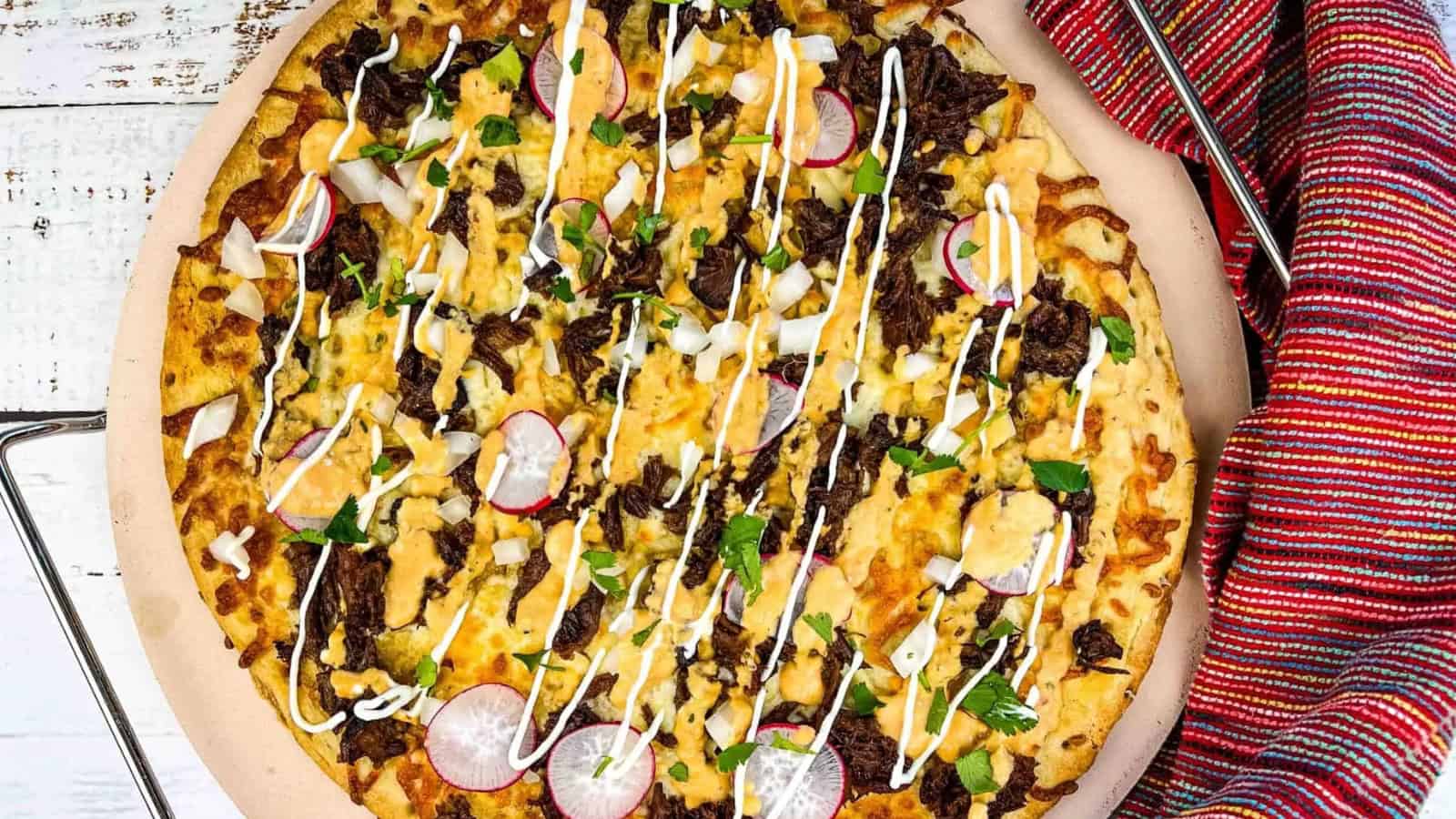 A Mexican-inspired pizza topped with beef, onions, radishes, cilantro, cheese, and drizzled with sour cream, on a wooden board next to a red-striped cloth.