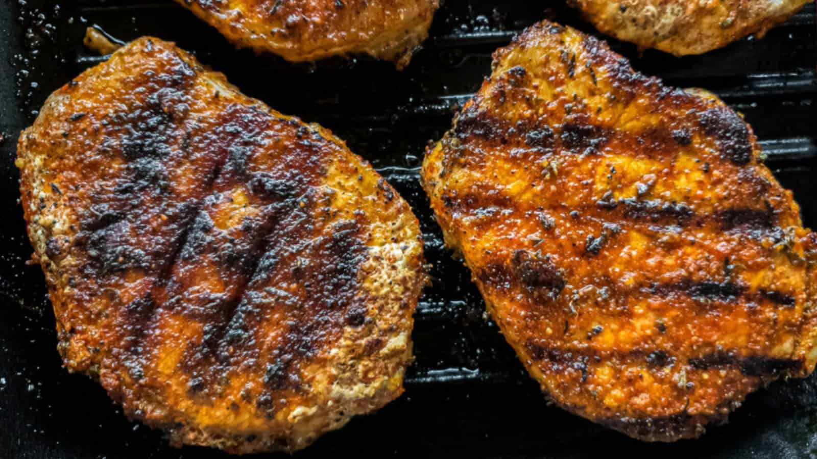 Close-up of two seasoned and grilled pork chops with distinct grill marks on a dark background.