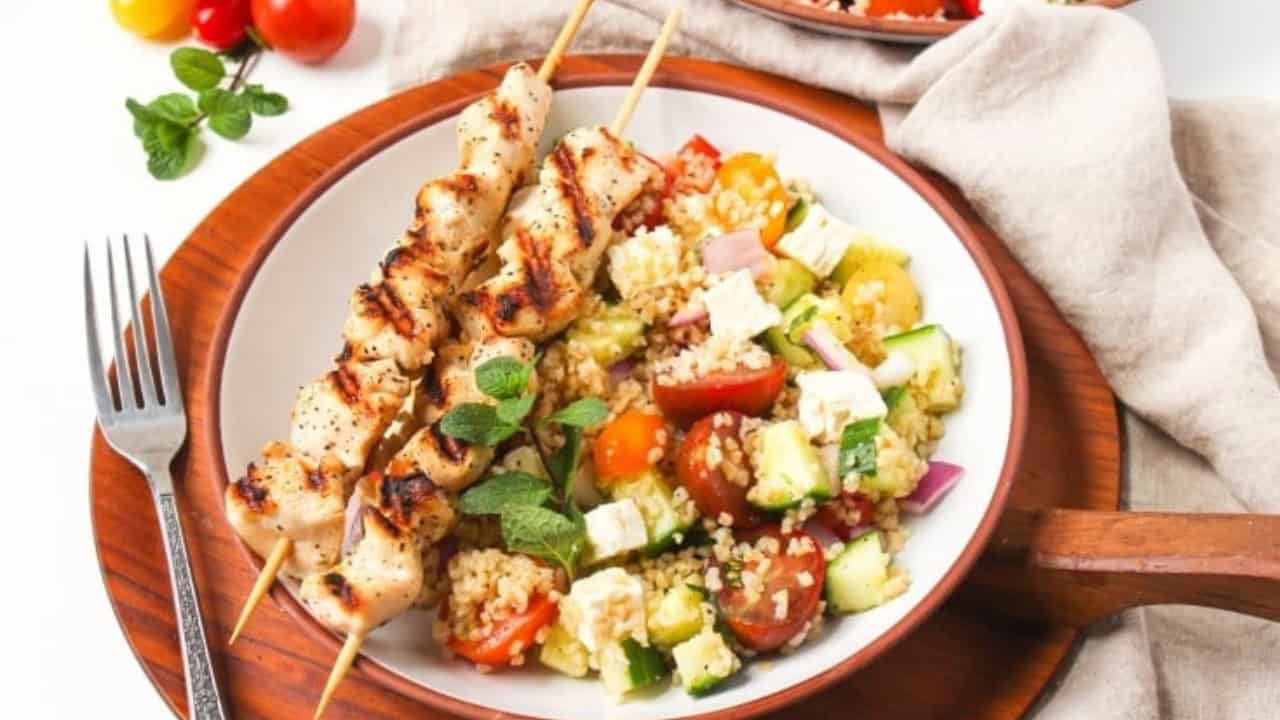 A plate with two grilled chicken skewers, bulgur salad with tomatoes, cucumber, feta, and mint. A fork is on the plate. Fresh tomatoes and mint are in the background.