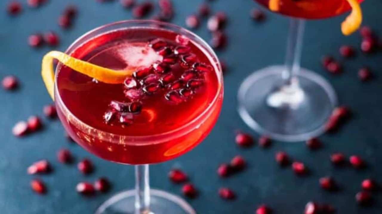 Two champagne pomegranate cocktails in coupe glasses on a dark blue background scattered with pomegranate seeds.