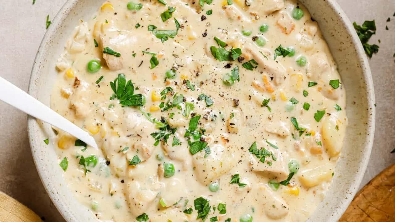 A creamy chicken and vegetable soup with peas, corn, and parsley garnished on top.