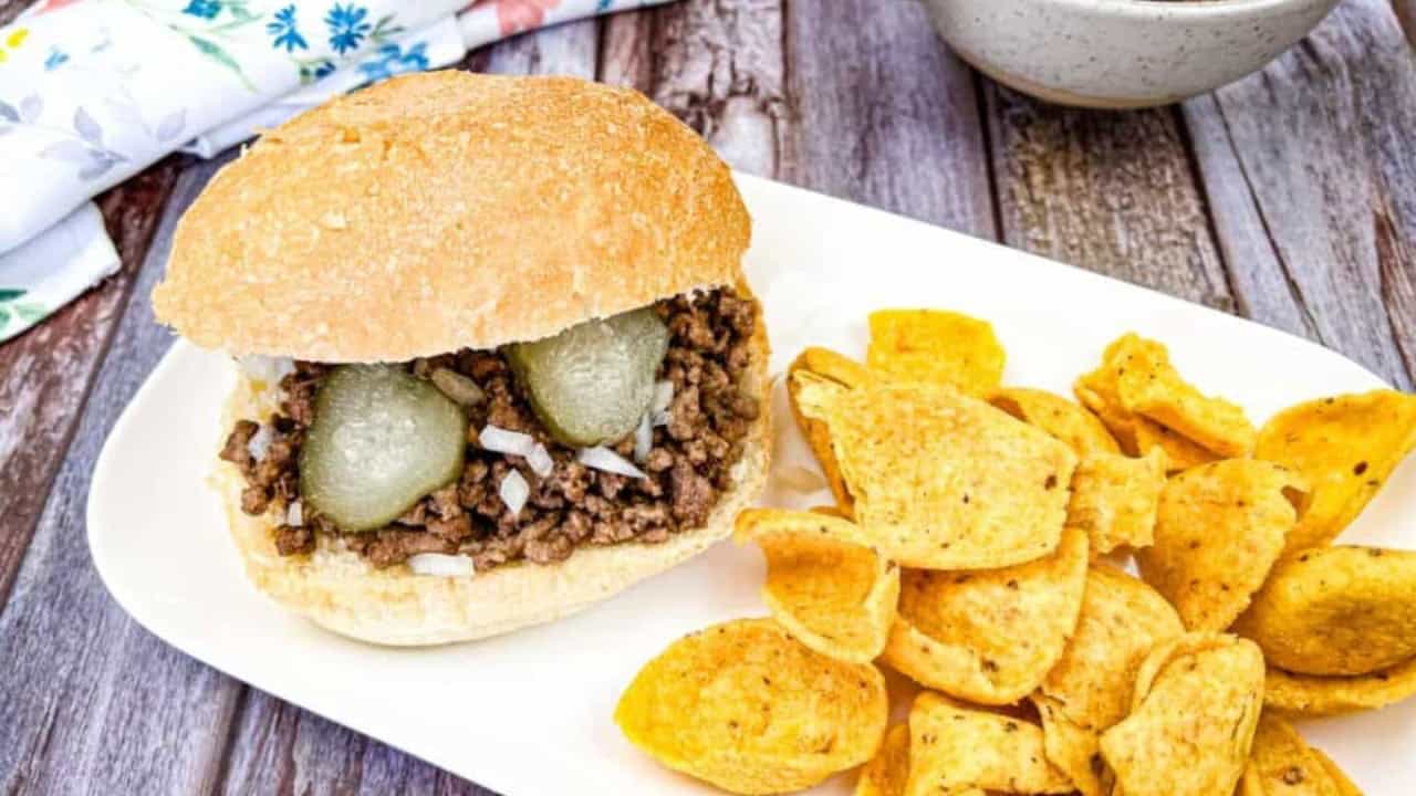 Sandwich with ground meat, pickles, and onions on a bun, served on a white plate with corn chips. A napkin and bowl are in the background.