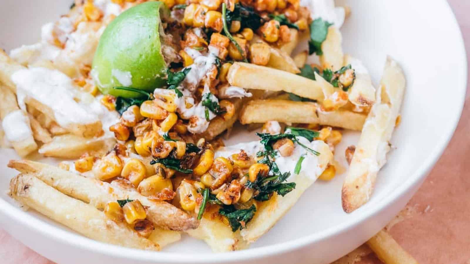A plate of fries topped with roasted corn, cilantro, and a lime wedge, drizzled with a white sauce.