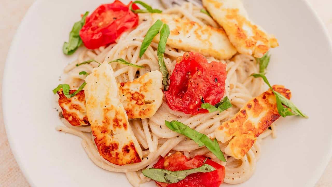 A plate of spaghetti topped with grilled halloumi cheese, sliced tomatoes, and fresh basil.