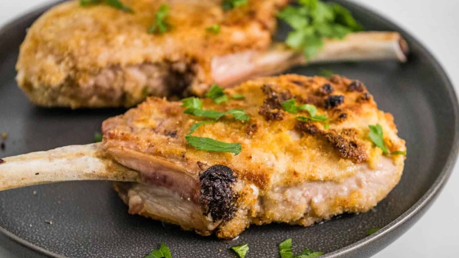 Two breaded and cooked pork chops on a dark plate, garnished with fresh parsley.