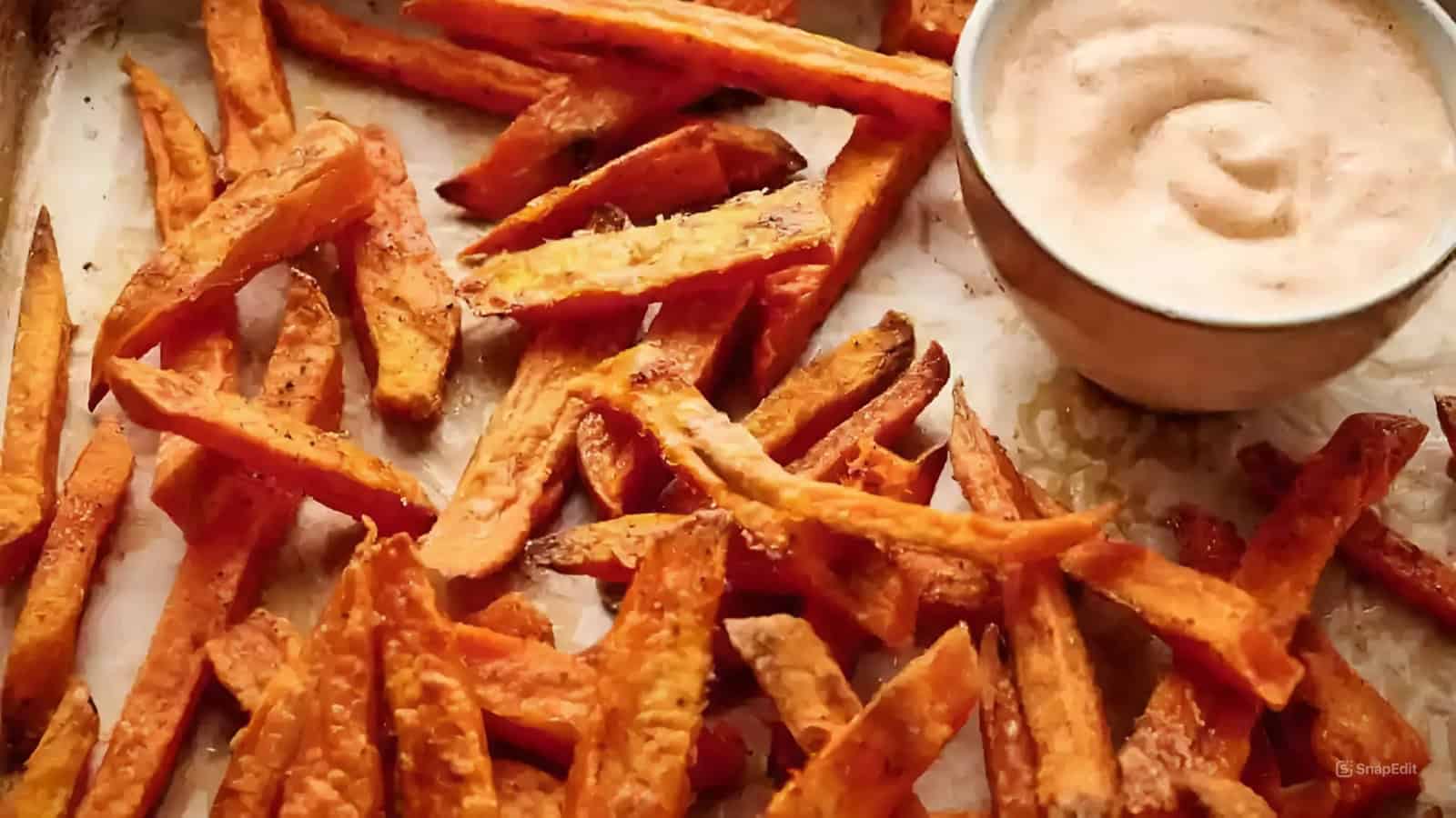 Sweet potato fries scattered on a tray with a bowl of creamy dipping sauce.