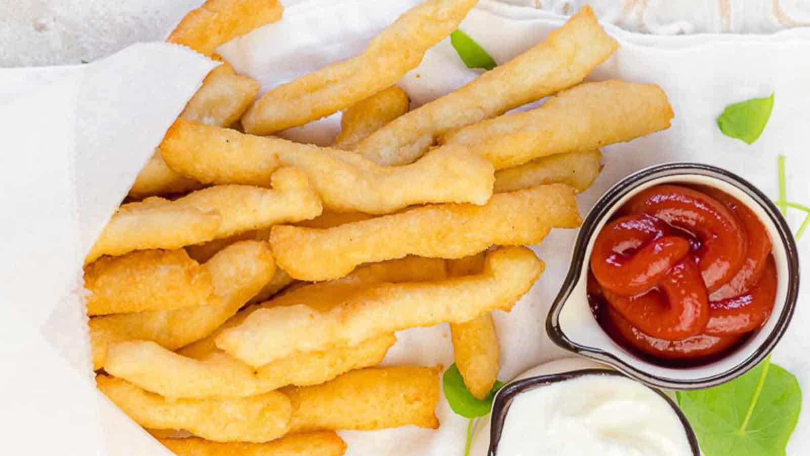 A pile of crispy fries in parchment paper next to small cups of ketchup and mayonnaise on a white surface.