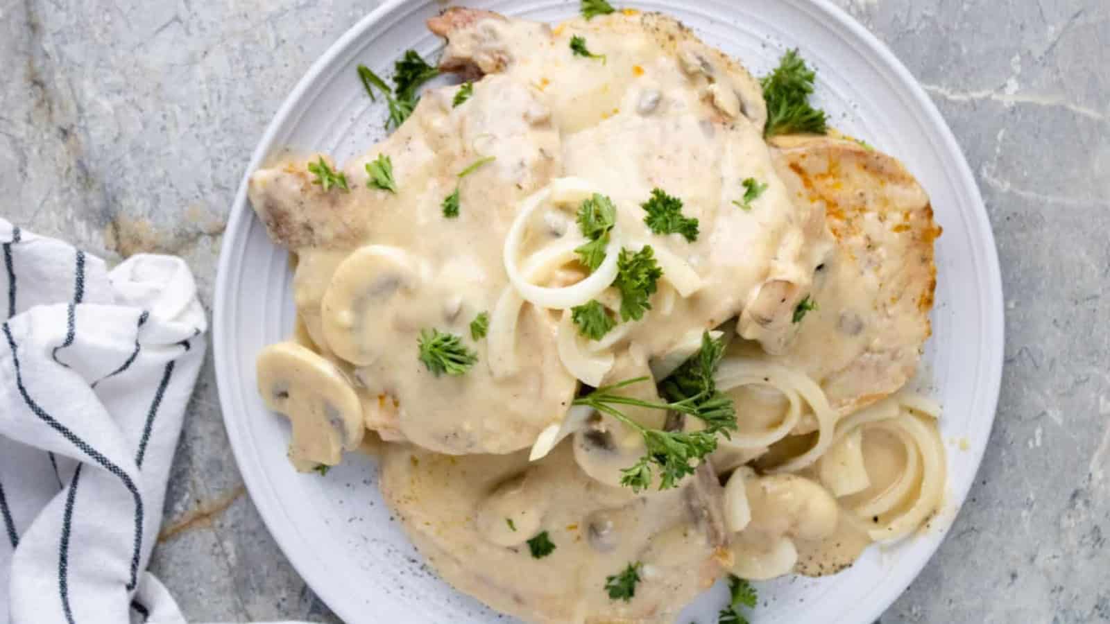 Plate of pork chops topped with creamy mushroom sauce, onion slices, and parsley garnish on a gray surface.