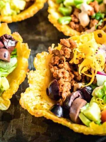 Colorful taco bowls filled with ground meat, diced tomatoes, lettuce, olives, cheese, and onions in crispy cheese shells on a dark plate.