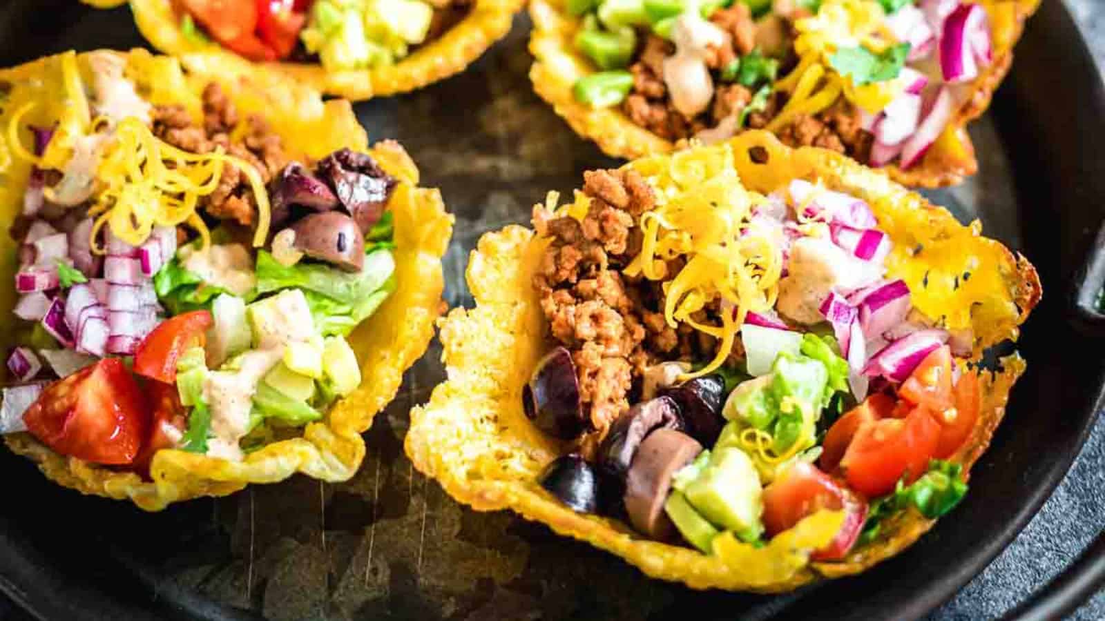 Colorful taco bowls filled with ground meat, diced tomatoes, lettuce, olives, cheese, and onions in crispy cheese shells on a dark plate.