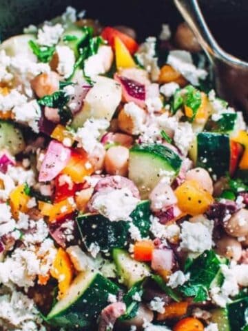 A bowl of colorful chopped salad with cucumbers, bell peppers, red onions, chickpeas, feta cheese, and herbs, served with a fork.
