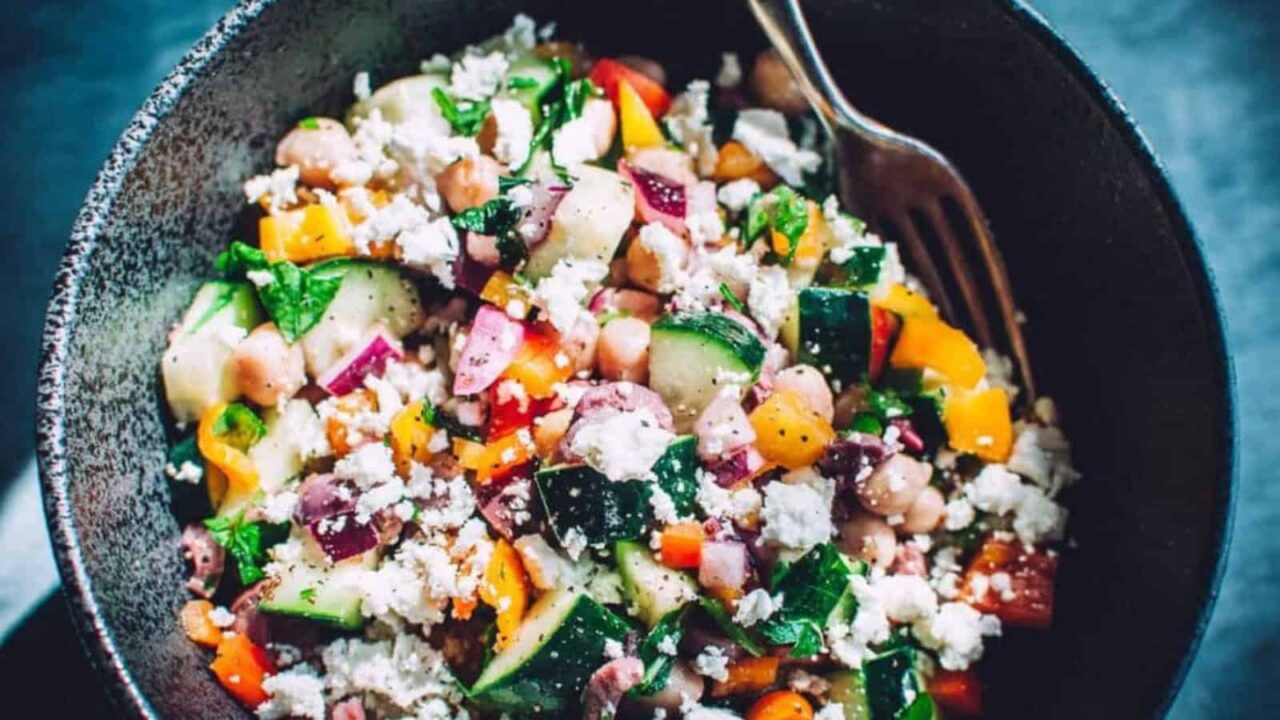A bowl of colorful chopped salad with cucumbers, bell peppers, red onions, chickpeas, feta cheese, and herbs, served with a fork.