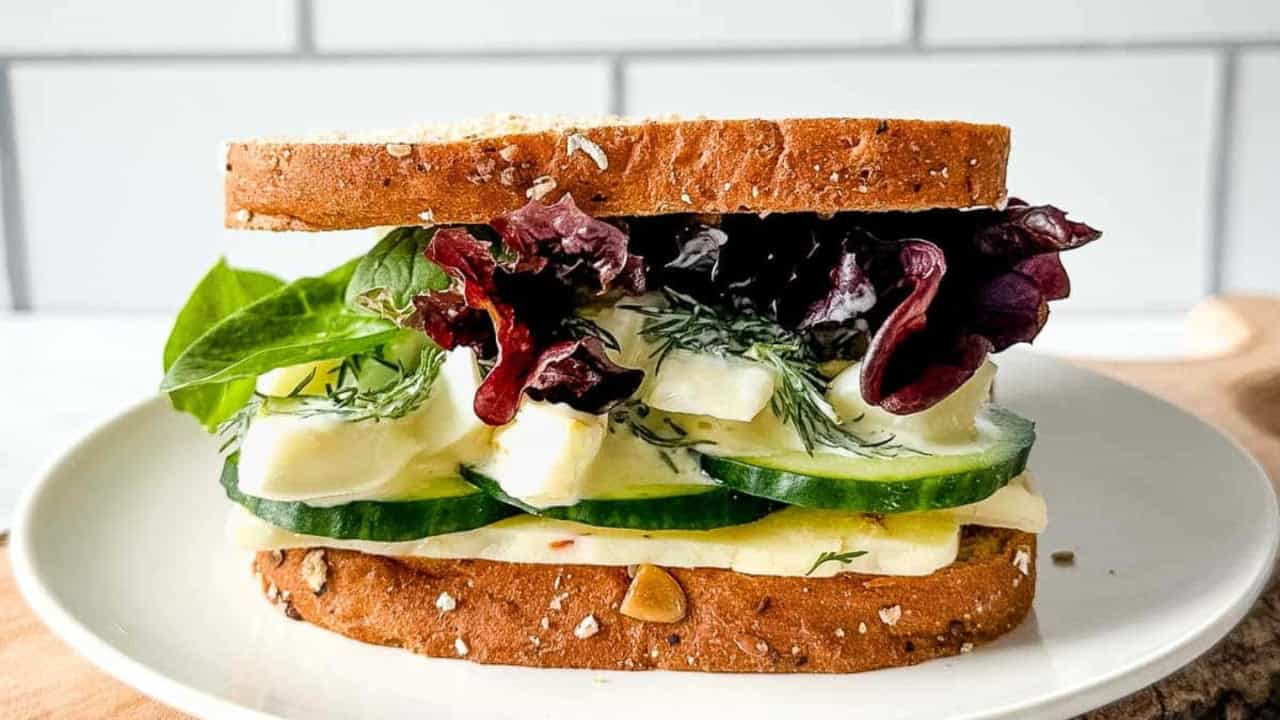 A sandwich with multigrain bread, lettuce, cucumber, cheese, and dill on a white plate against a tiled background.