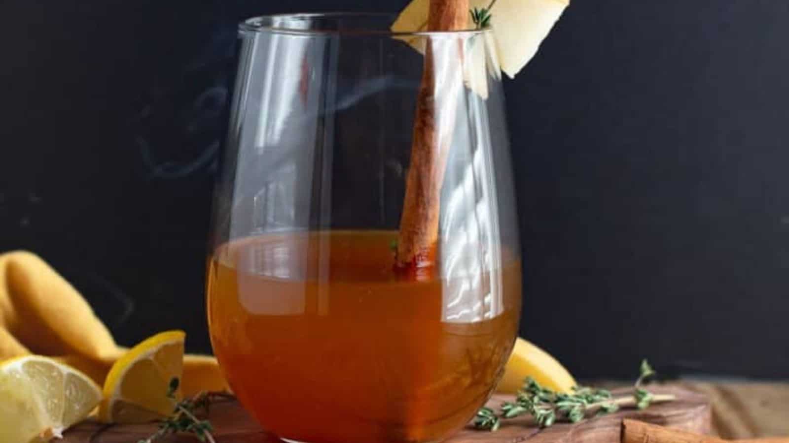A glass of warm cider garnished with a cinnamon stick and apple slice, placed on a wooden surface with assorted lemons and leaves surrounding it.