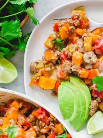 A plate of colorful beef and vegetable stir-fry with avocado slices and lime wedges on the side, garnished with cilantro.