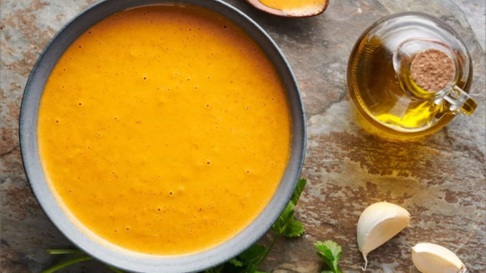 A bowl of orange soup garnished with herbs is placed on a stone surface. Nearby, there's a halved mango, a sprig of cilantro, two garlic cloves, and a glass bottle of oil.