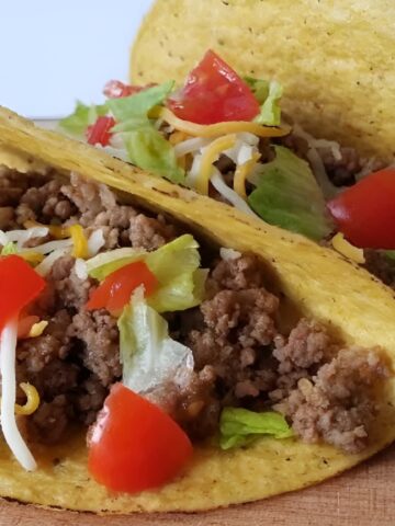Three hard shell tacos filled with ground beef, diced tomatoes, shredded lettuce, and cheese, placed on a wooden surface.