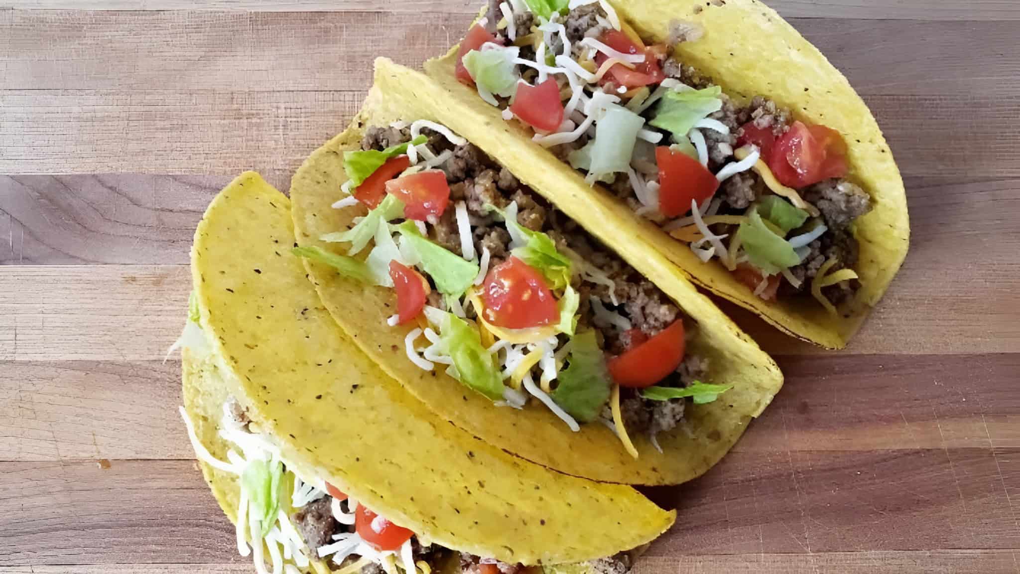 Three hard shell tacos filled with ground beef, shredded cheese, lettuce, and diced tomatoes on a wooden surface.