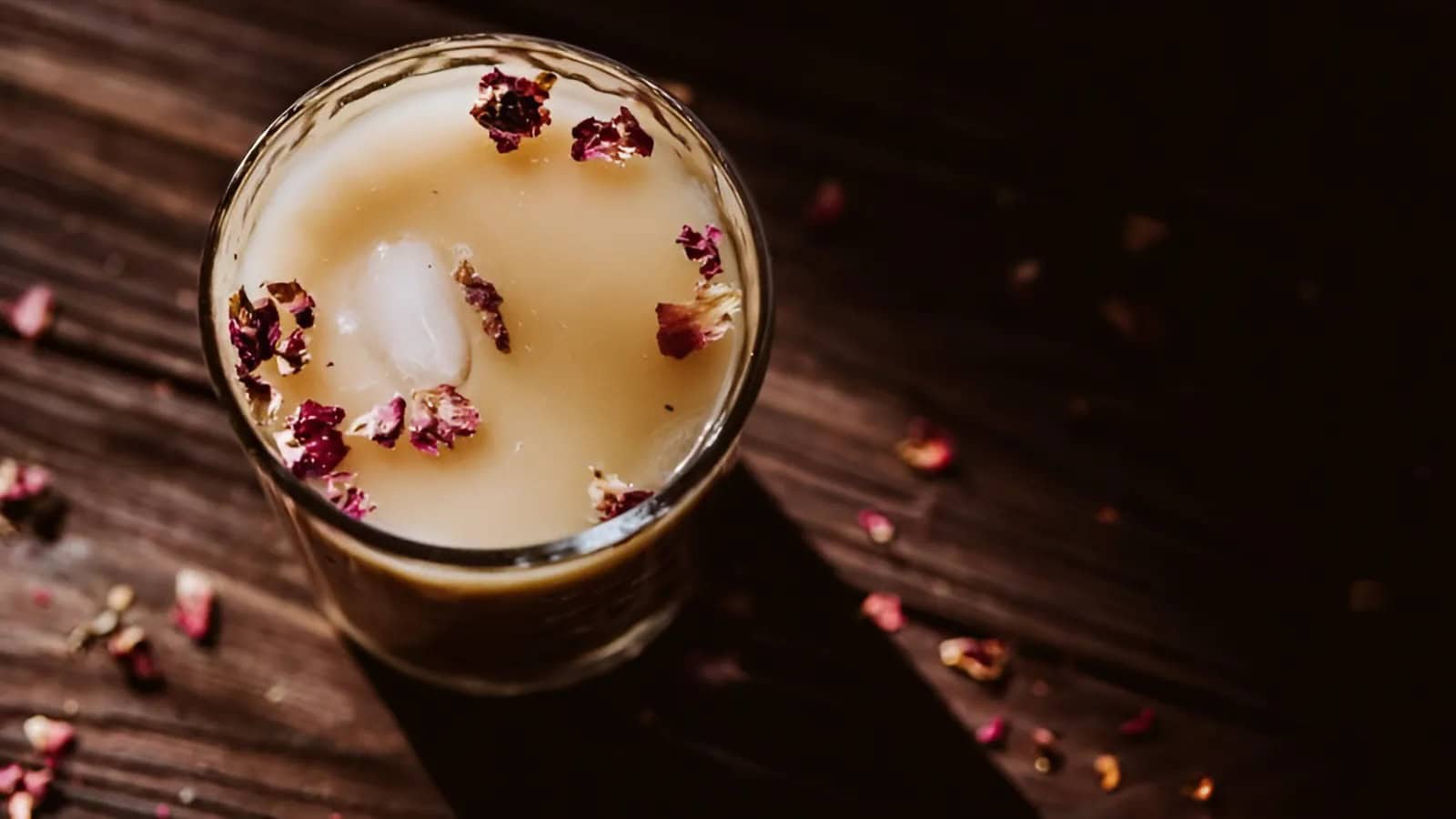 A glass of creamy iced chai tea is topped with rose petals, sitting on a wooden surface.