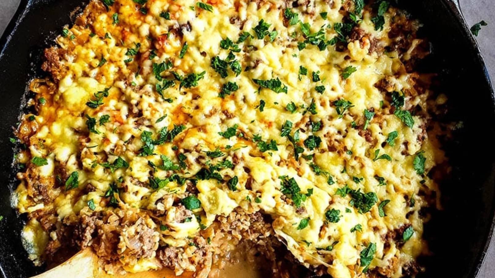 A skillet filled with baked ground meat, rice, and melted cheese, garnished with chopped herbs. A portion is missing, revealing layers underneath.