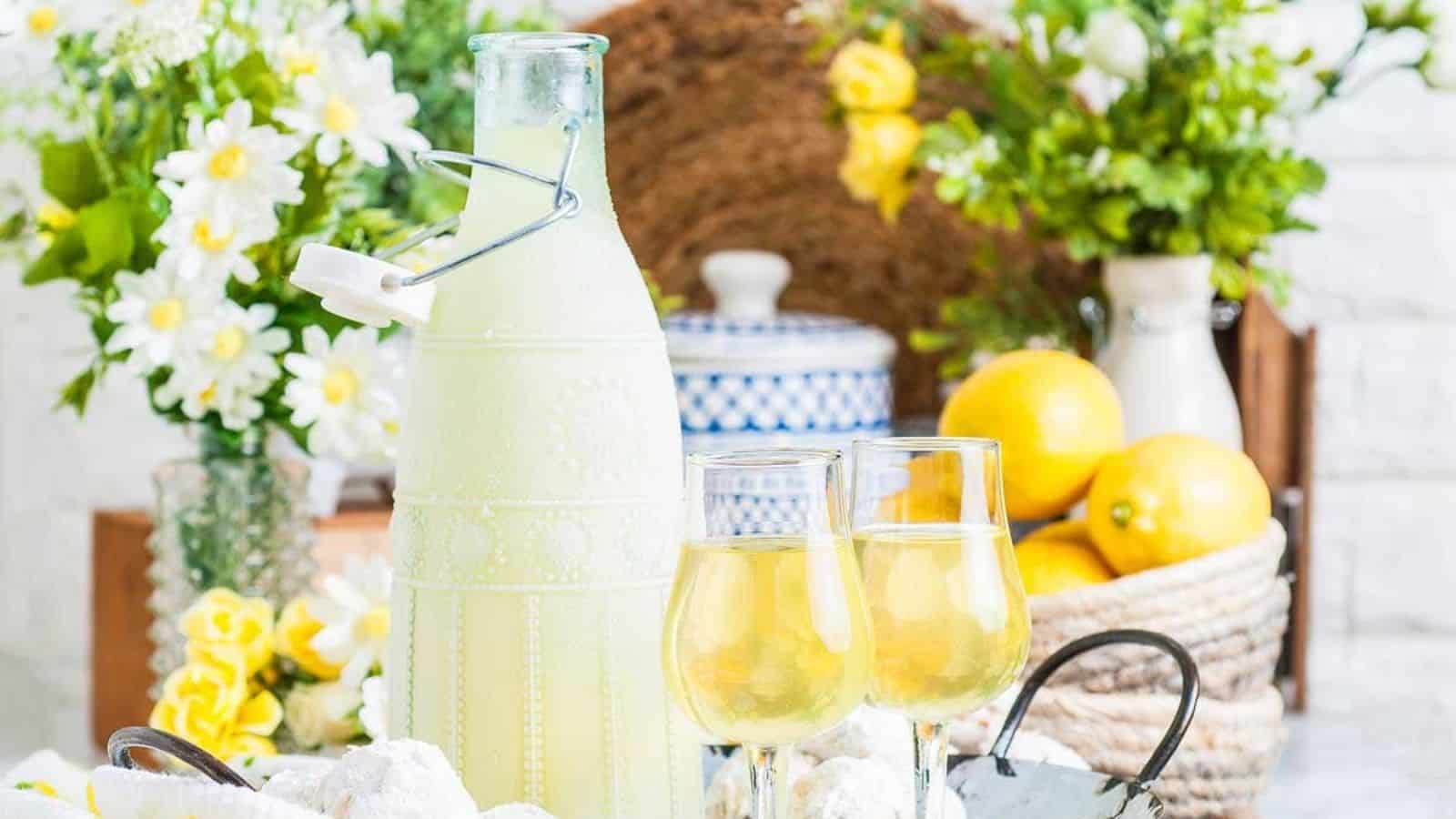 A bottle and two glasses of lemonade are on a table with flowers and a basket of lemons in the background.