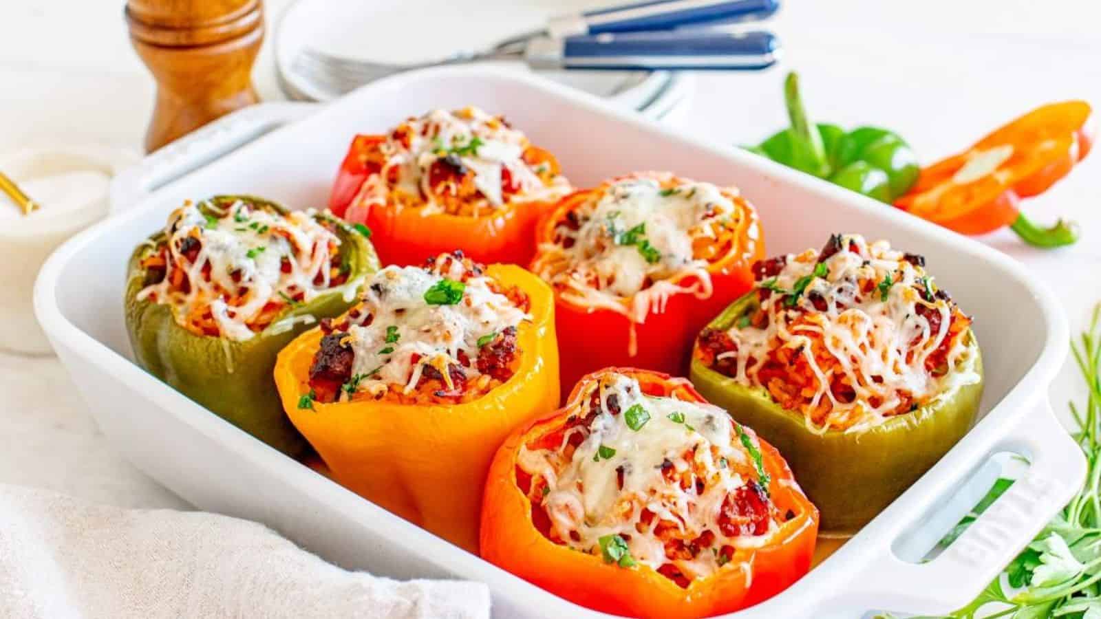 A white baking dish contains six stuffed bell peppers topped with melted cheese and herbs. Peppers are red, yellow, and green. Plates and utensils are in the background.