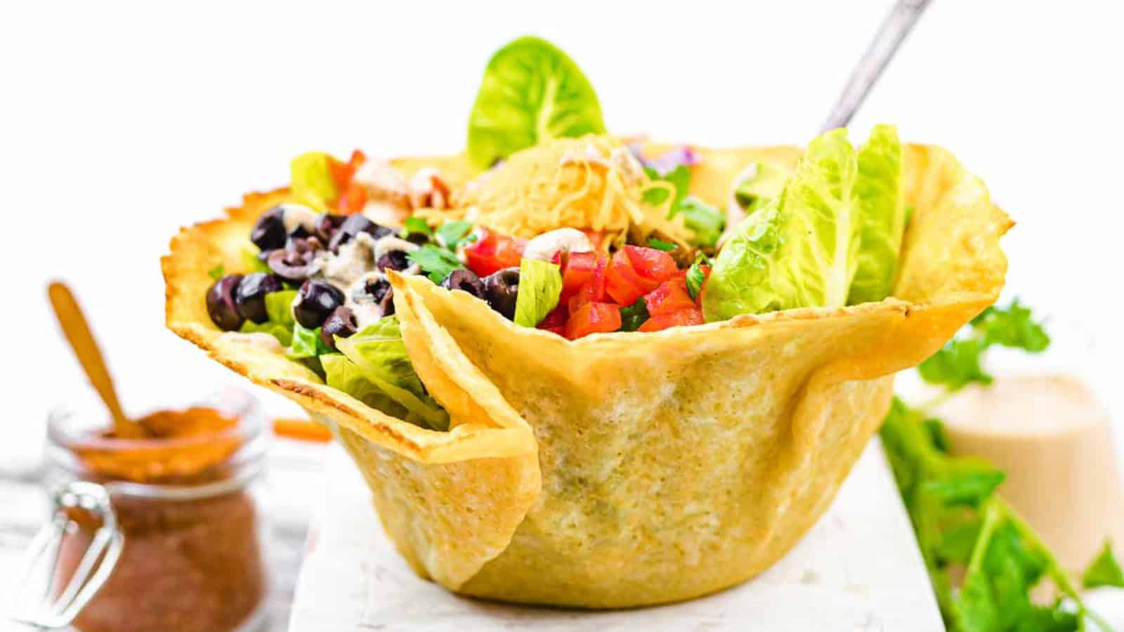 Taco salad in a tortilla bowl, topped with lettuce, tomatoes, black olives, shredded cheese, and a dollop of sour cream. A jar of seasoning is in the background.