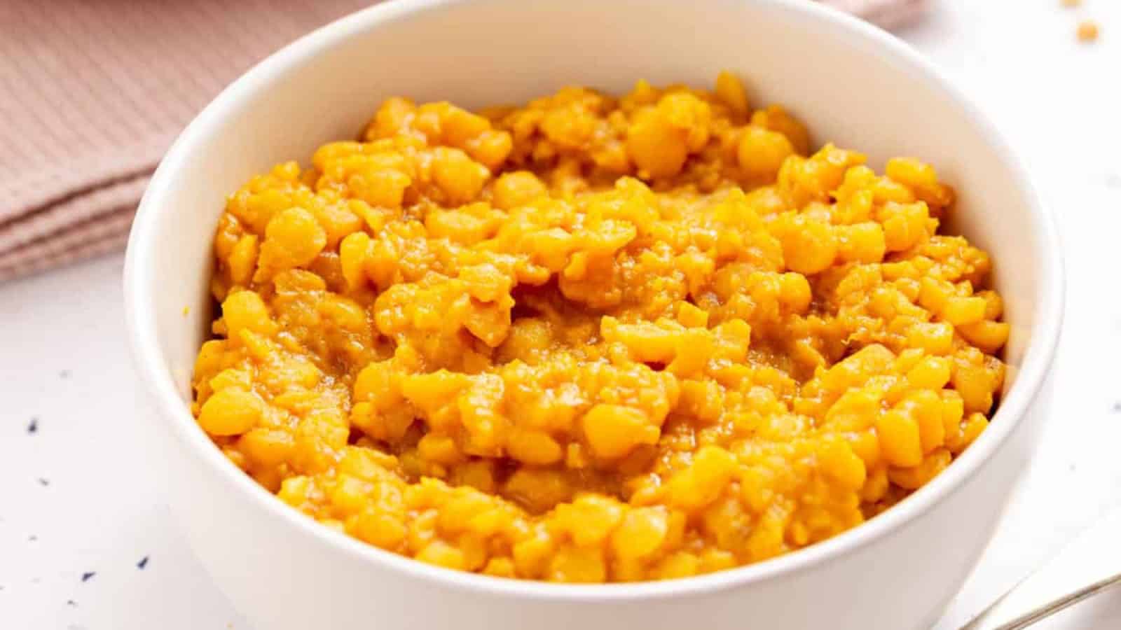 A white bowl filled with cooked yellow split peas on a light surface with a pink napkin in the background.