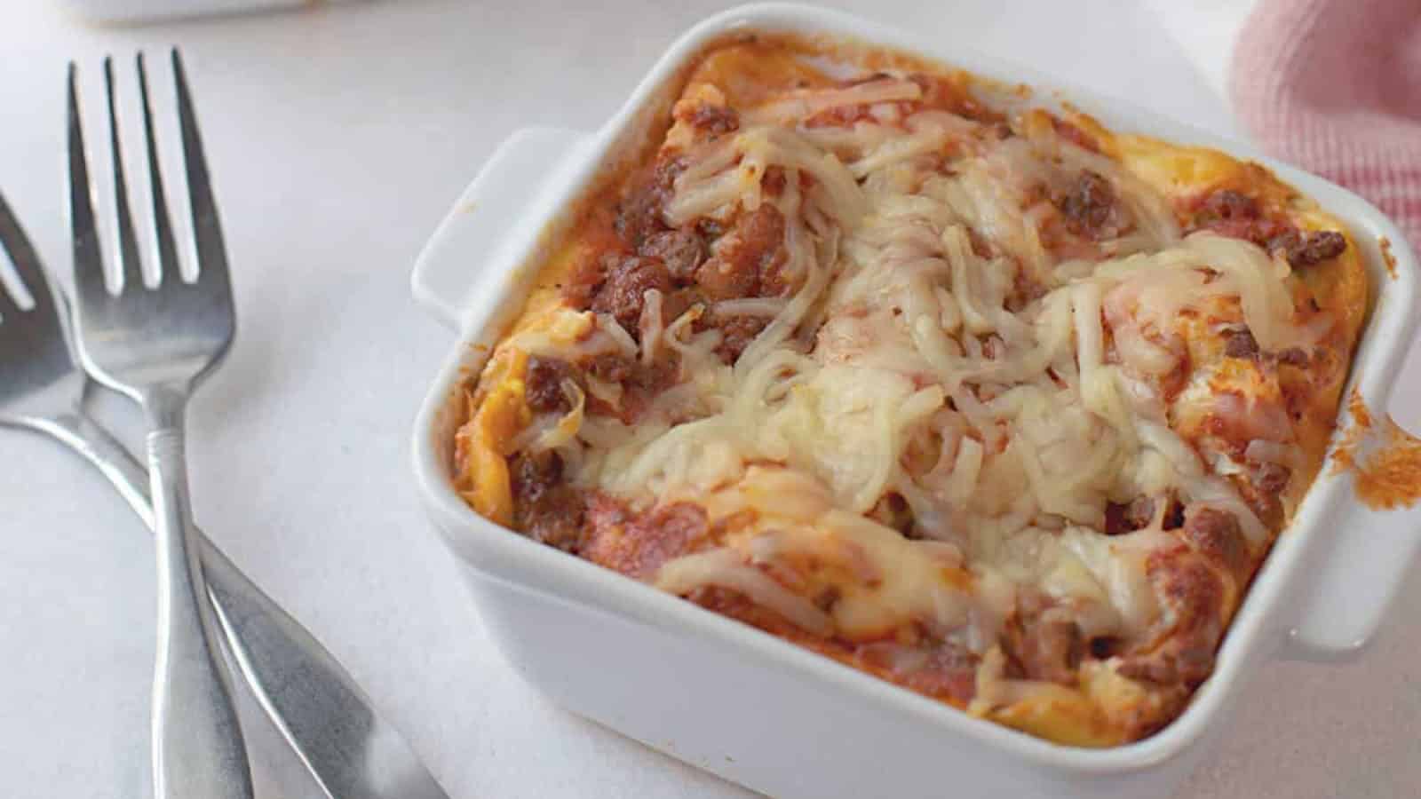 A square dish of baked lasagna topped with melted cheese is placed on a table next to three forks.