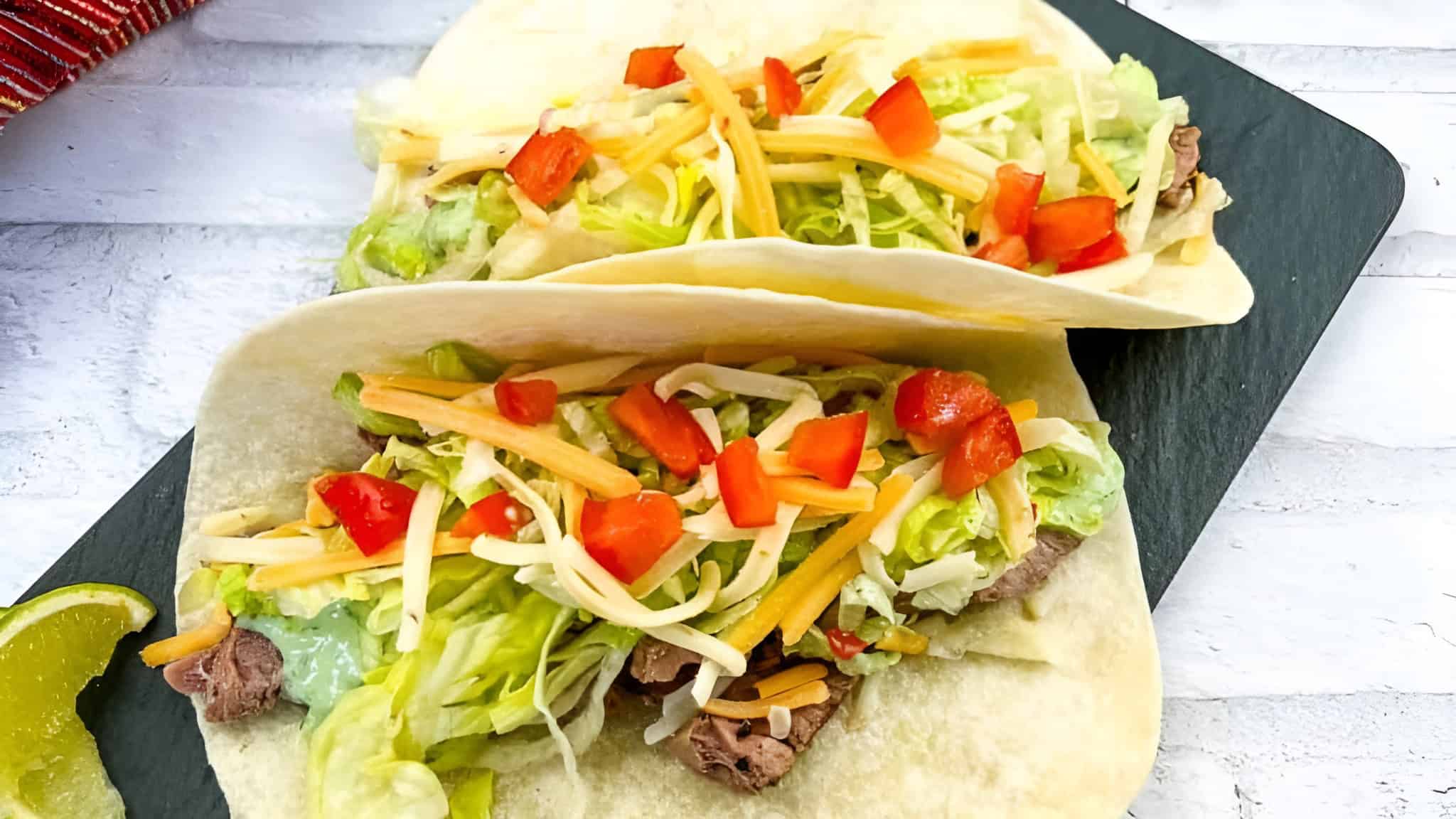 Two soft tacos filled with lettuce, shredded cheese, diced tomatoes, and beef on a slate board, with a lime wedge on the side.