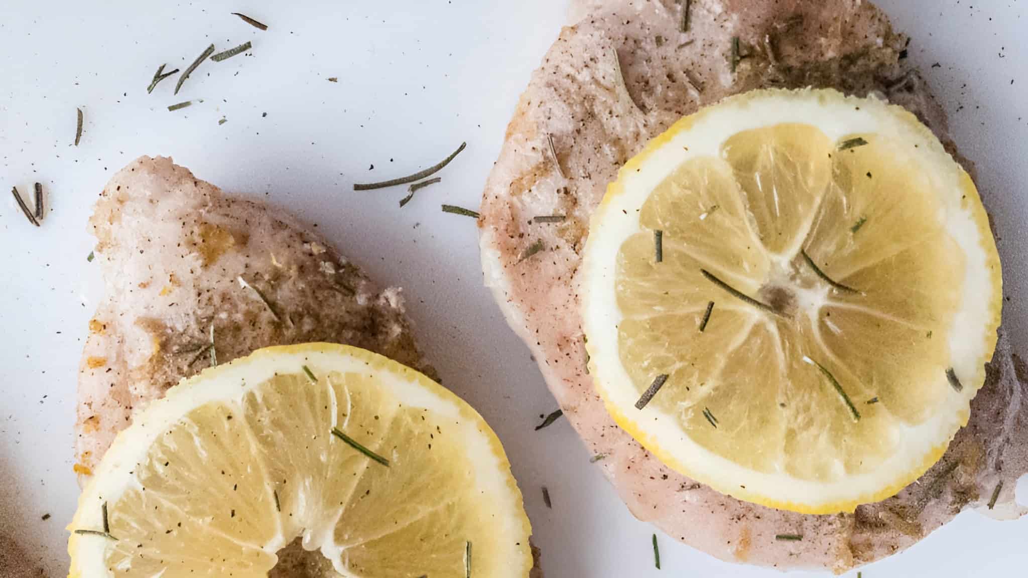 Two seasoned chicken breasts topped with lemon slices and herbs on a white surface.