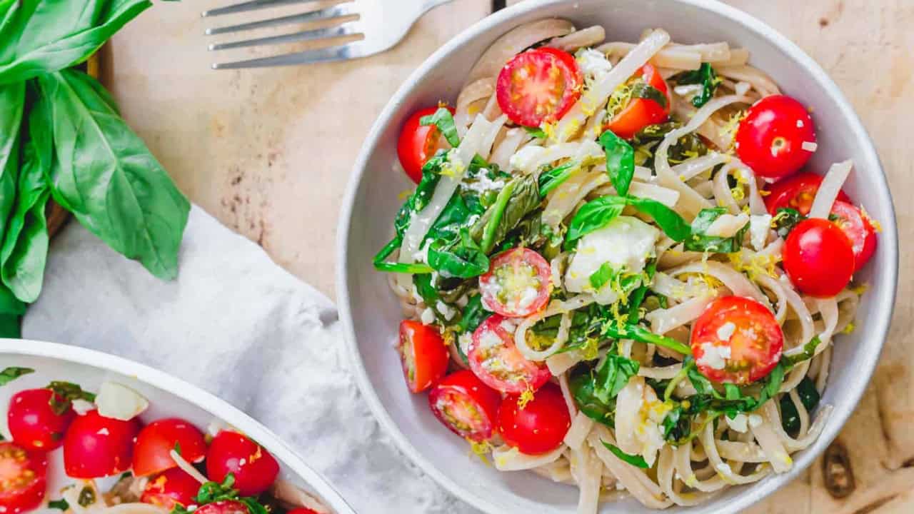 A bowl of pasta with cherry tomatoes, spinach, and lemon zest on a table with a fork and basil leaves.
