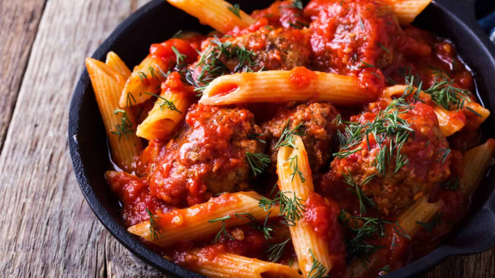 A skillet filled with penne pasta, meatballs, and tomato sauce, garnished with fresh dill, placed on a wooden surface.