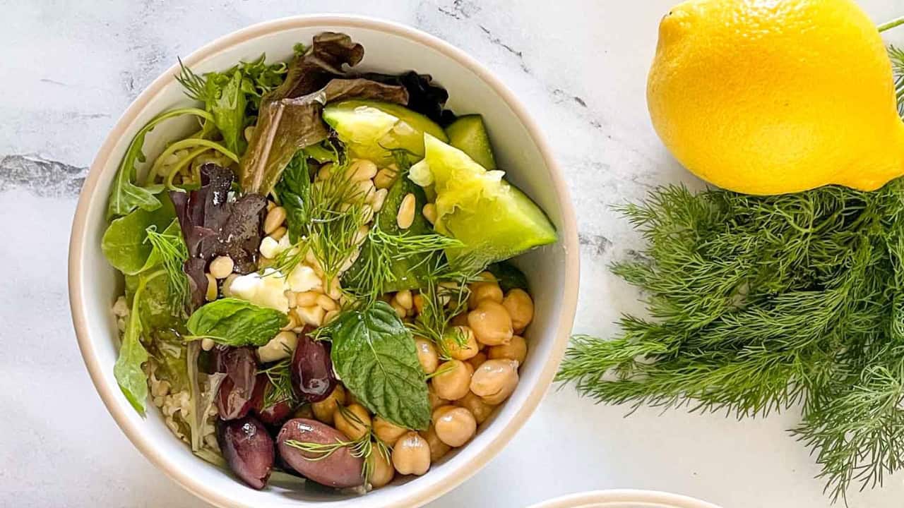 A bowl of mixed greens, chickpeas, cucumber, kalamata olives, pine nuts, and herbs, next to a lemon and dill bunch on a marble surface.