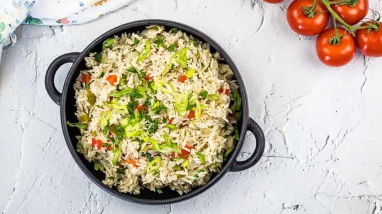 A black bowl filled with rice mixed with chopped vegetables and herbs, placed on a textured white surface, with a few red tomatoes beside it.