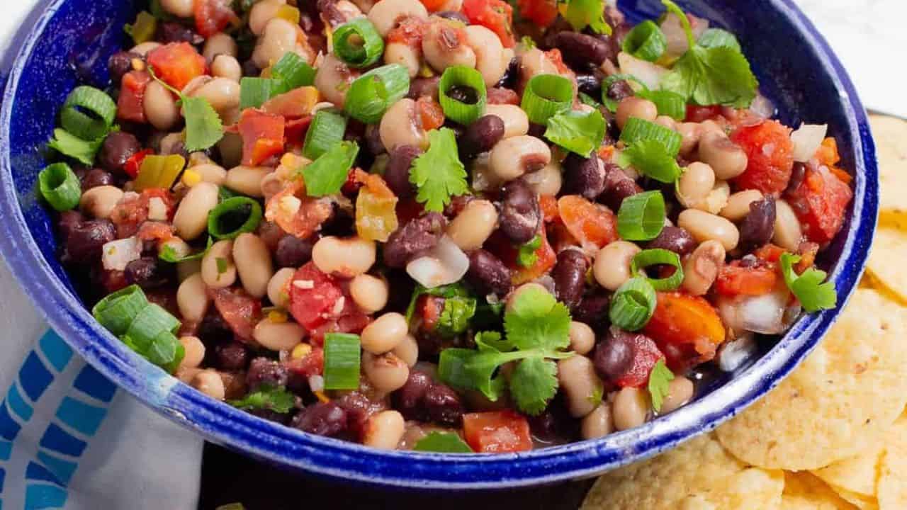 A blue bowl filled with a colorful bean salad, featuring black beans, diced tomatoes, onions, green onions, and cilantro, next to a pile of tortilla chips.