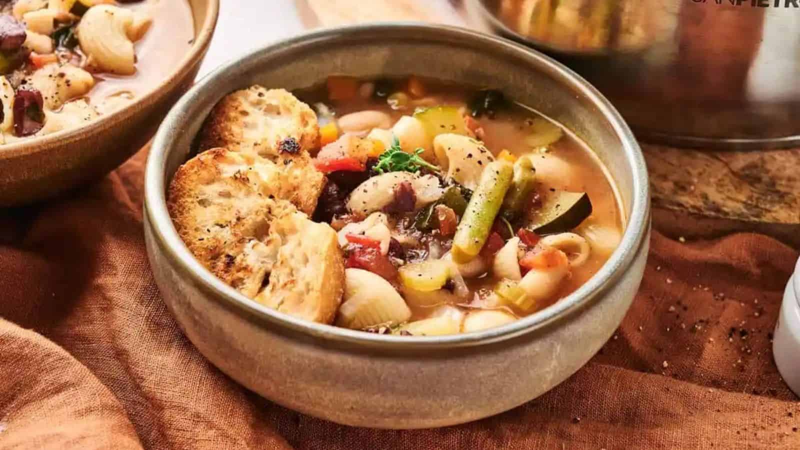 A bowl of vegetable soup with pasta and beans, garnished with a toasted bread slice.