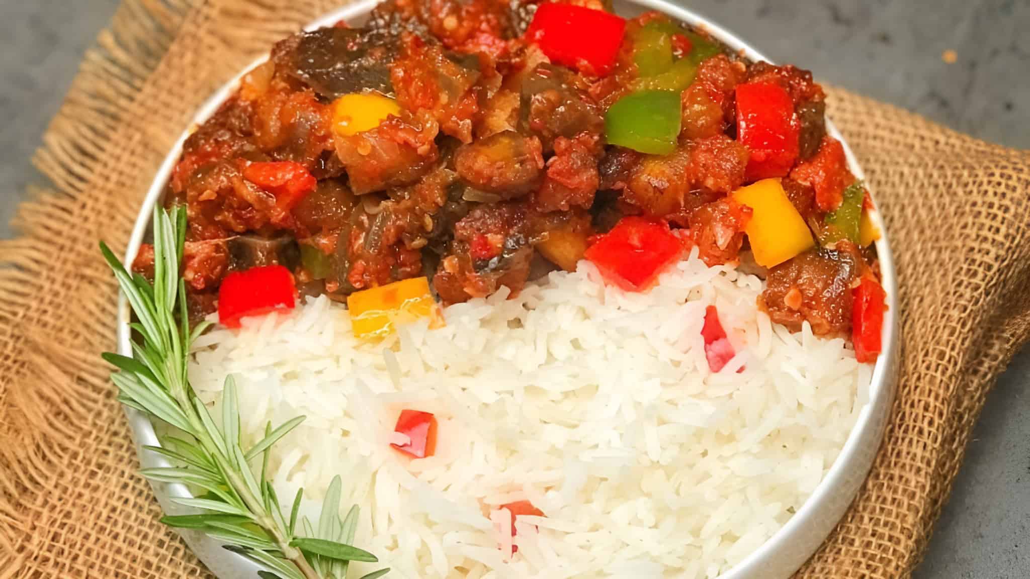 A plate of white rice topped with a colorful vegetable stew, garnished with a sprig of rosemary, on a burlap cloth.