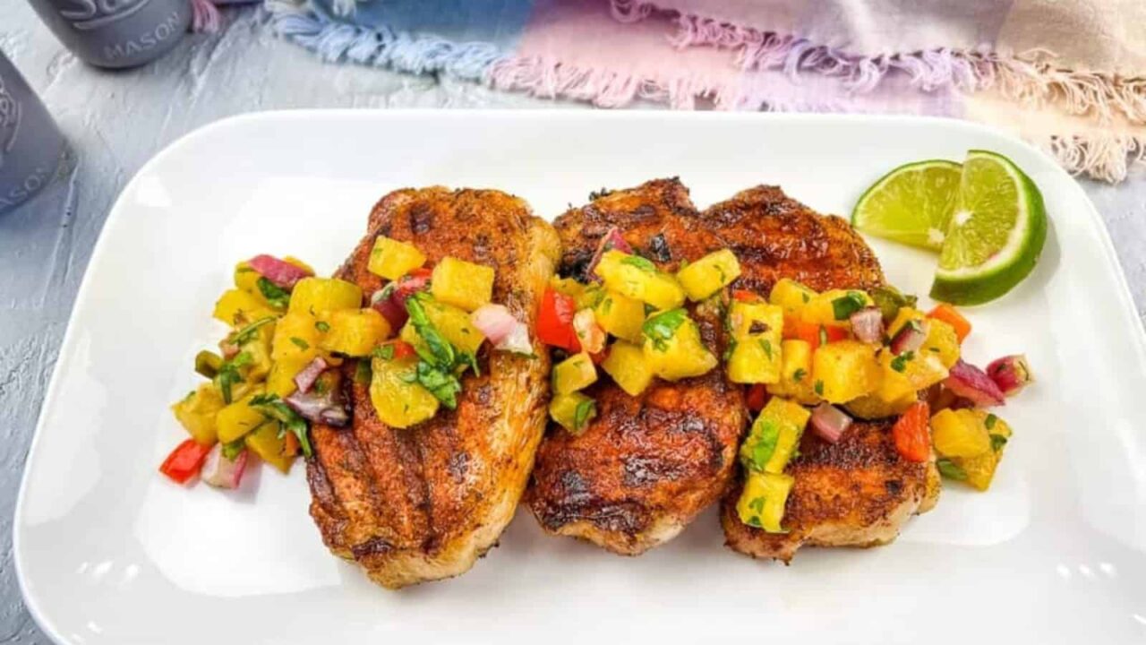 Three grilled pork chops topped with colorful pineapple salsa and a lime wedge on a white plate.