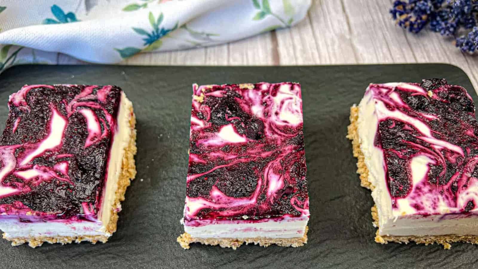 Three cheesecake bars with a marbled blueberry topping on a dark rectangular slate board.