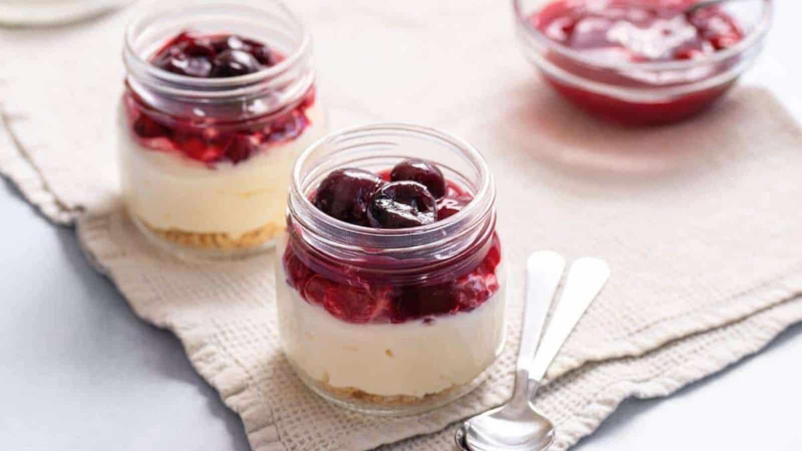 Two jars of cheesecake topped with cherry sauce on a beige cloth, with a small bowl of cherries and two spoons nearby.