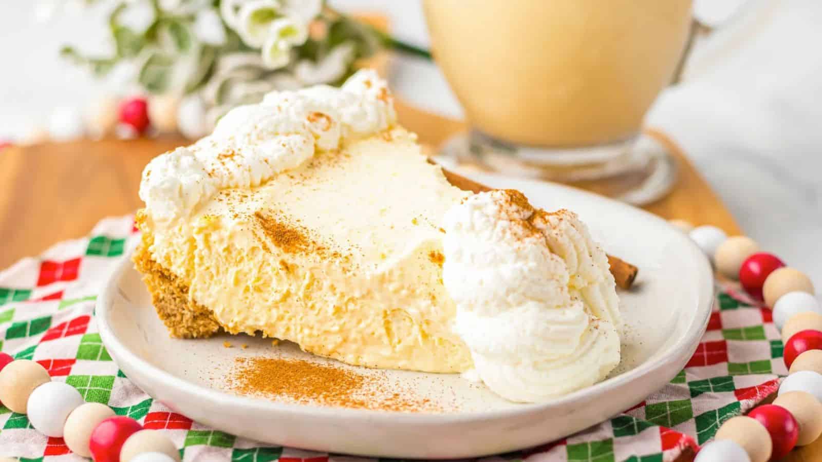 A slice of creamy pie with whipped cream sits on a white plate, garnished with cinnamon. A mug is in the background, and the plate is on a colorful, checkered cloth.