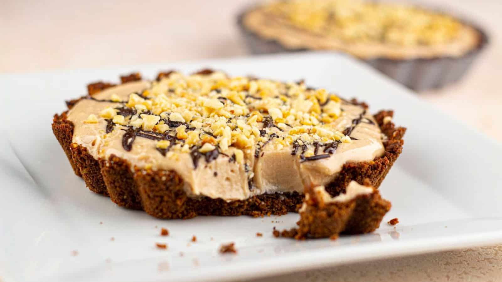 A small pie on a white plate with a chocolate crust, cream filling, and chopped nuts on top. Another pie is blurred in the background. A piece is missing from the front.