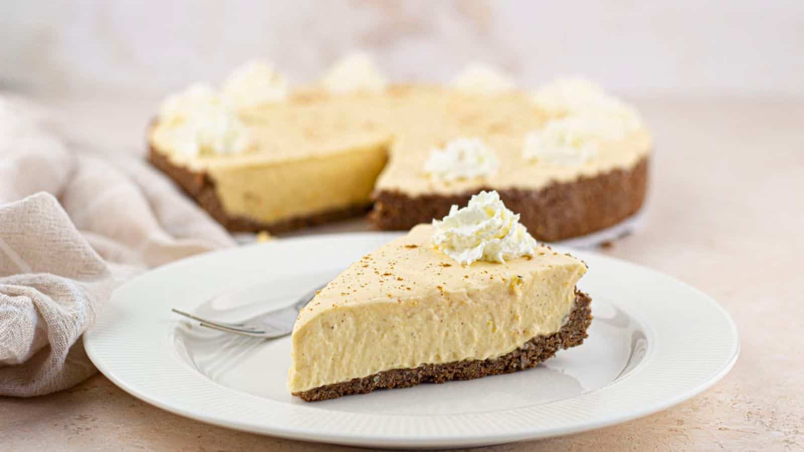 A slice of creamy pie with a crumbly crust is on a white plate, topped with whipped cream, with the rest of the pie in the background. A fork rests beside the slice.
