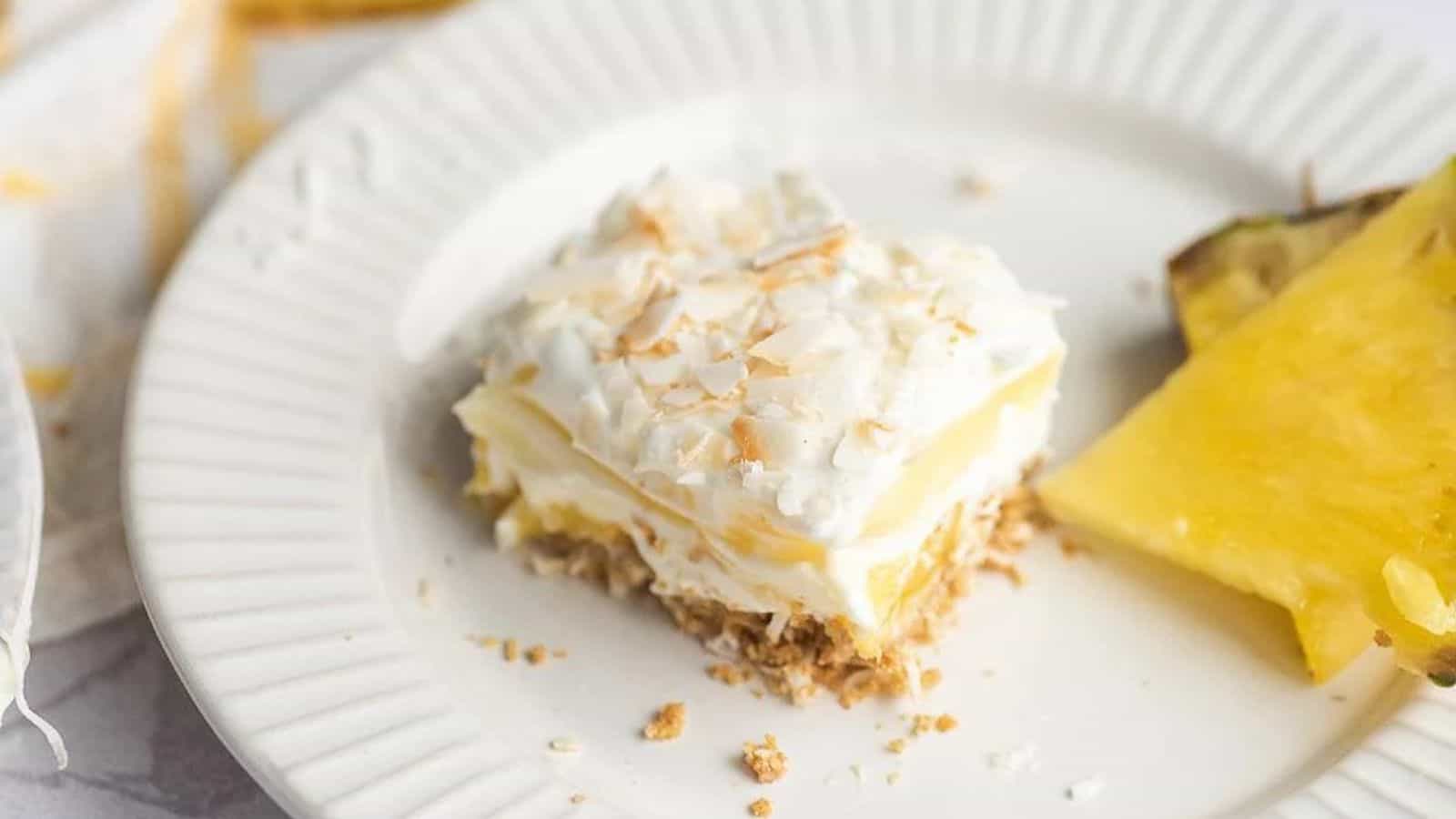 A slice of coconut cream pie with a toasted coconut topping on a white plate, accompanied by a wedge of pineapple.